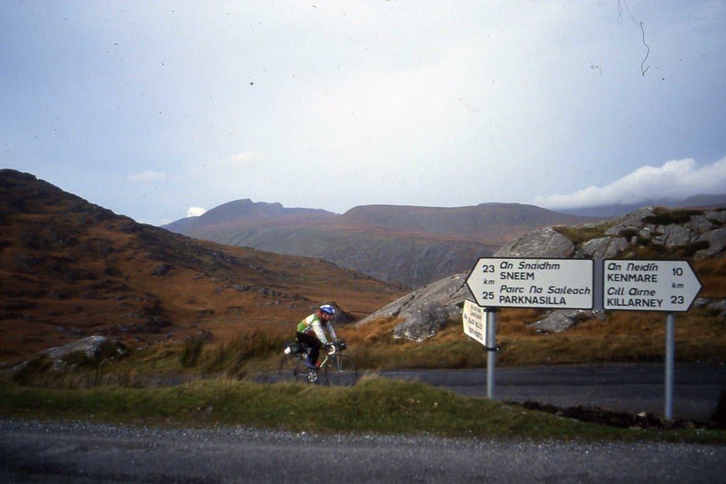 A cyclist rides along the Ring of Kerry past road signs pointing toward Sneem, Kenmare, and Killarney against a backdrop of rolling mountains.
