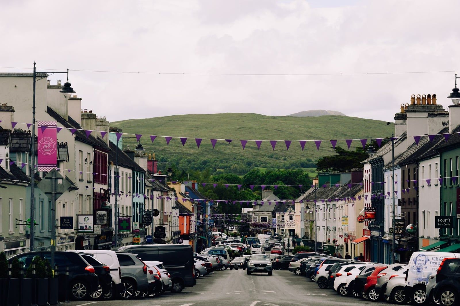 Parked cars line a colourful street adorned with purple bunting and flanked by shops in Kenmare on the Ring of Kerry.