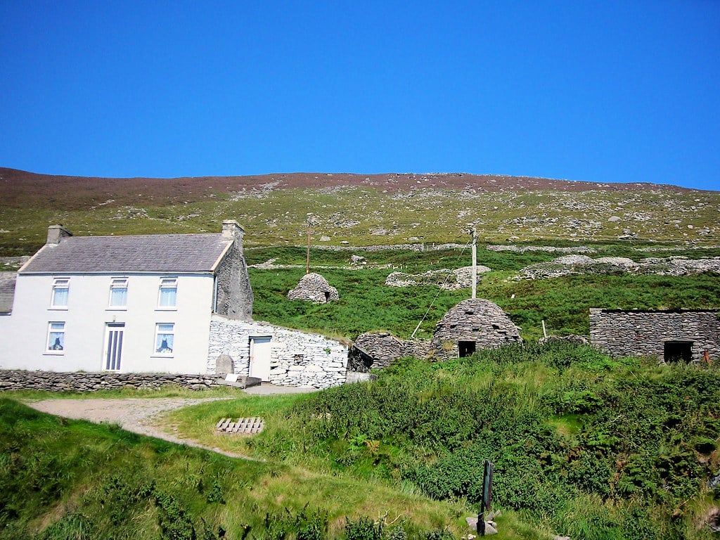 Tradizionali capanne di pietra a forma di alveare si ergono lungo un fianco collinare erboso accanto a un cottage imbiancato a calce sulla Penisola di Dingle.
