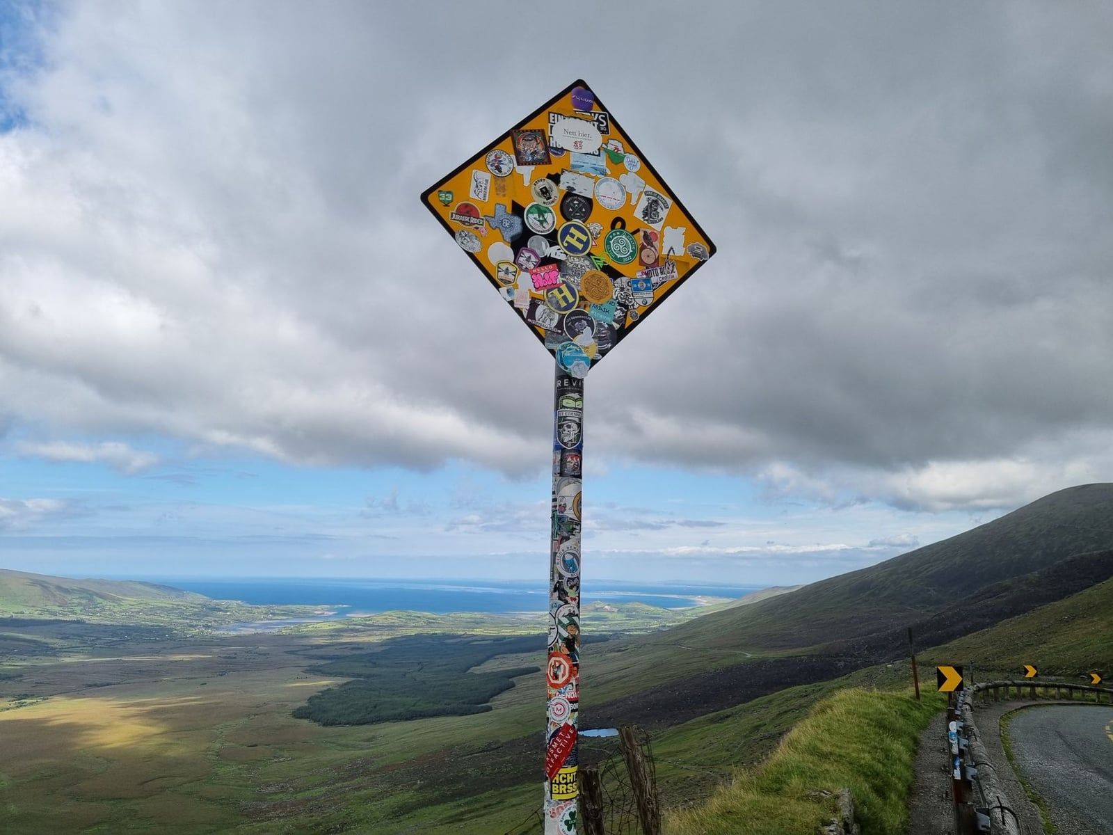 Un cartello stradale coperto di adesivi si affaccia su una strada di montagna tortuosa e sulla valle al Conor Pass sulla Penisola di Dingle.