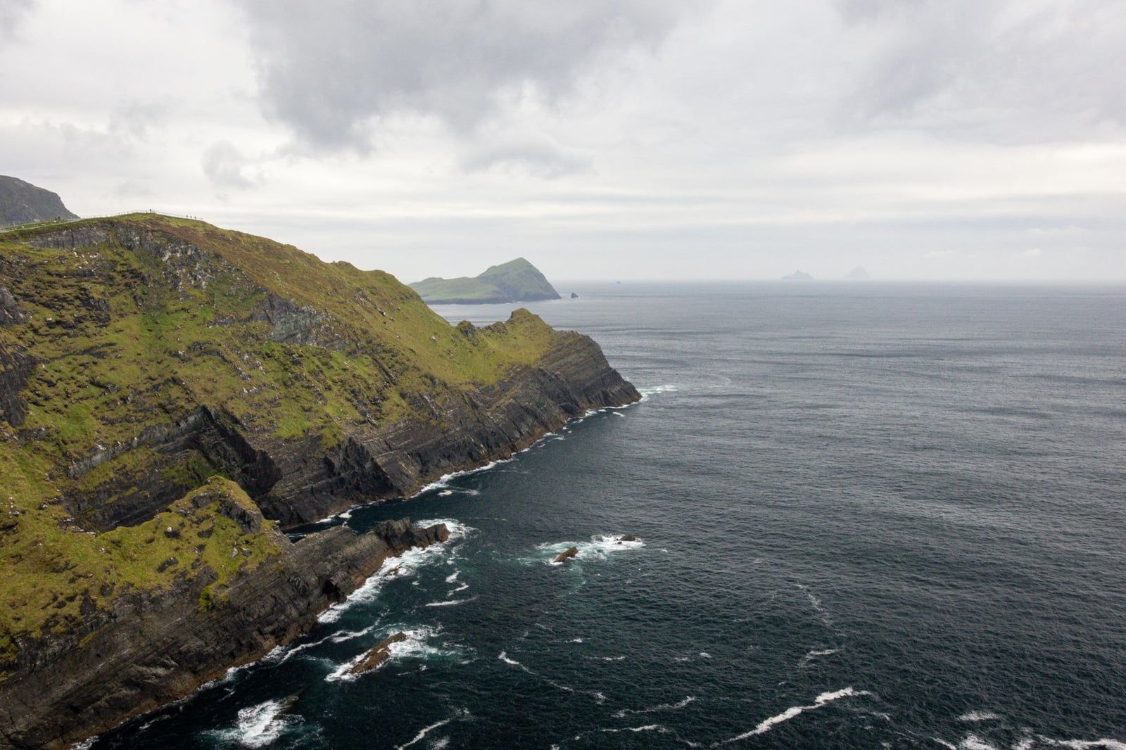 Rugged sea cliffs plunge into the Atlantic Ocean with Puffin Island and the Skellig Islands visible on the horizon along the Ring of Kerry.